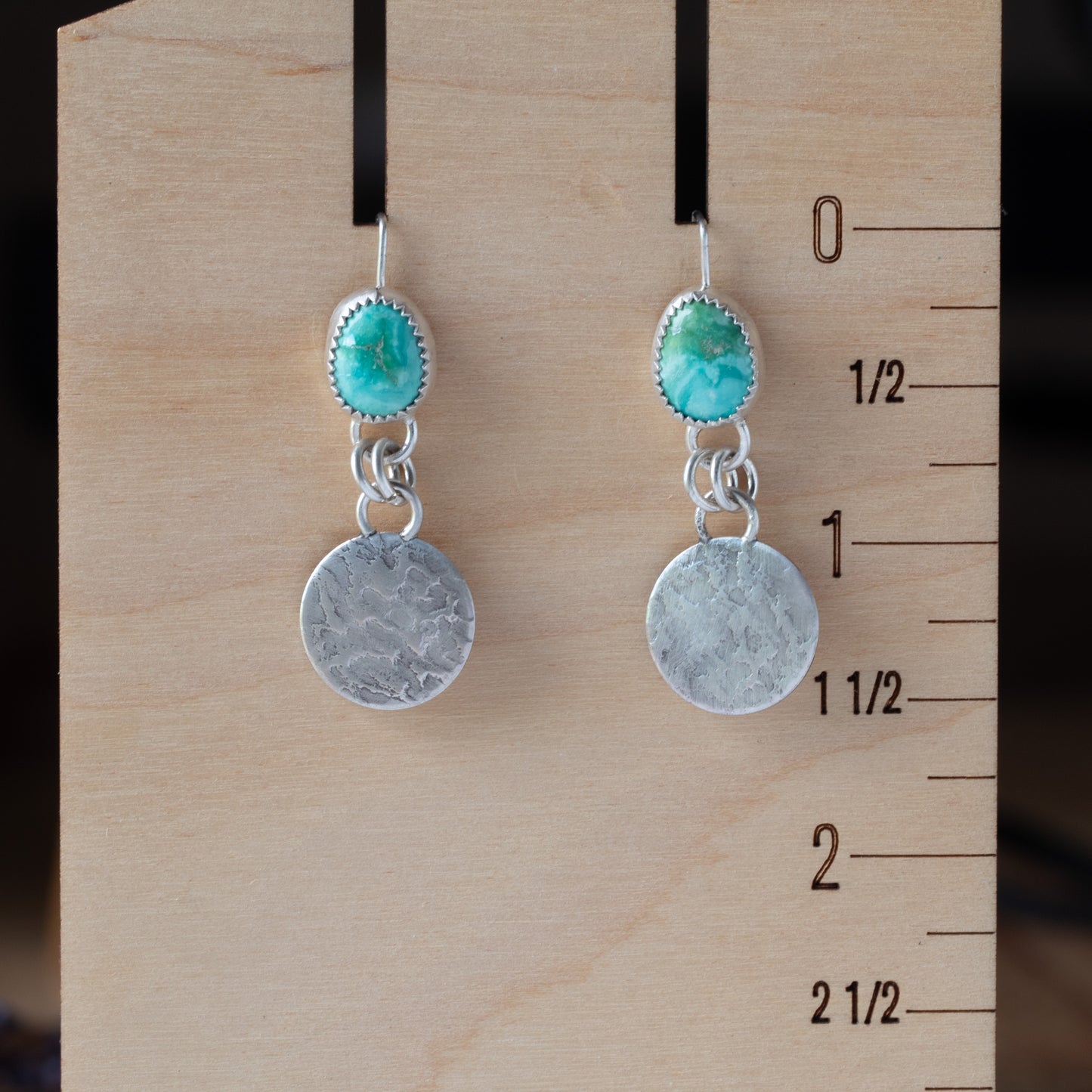 Pair of earrings with turquoise stones and silver discs on a wooden block with measurements.