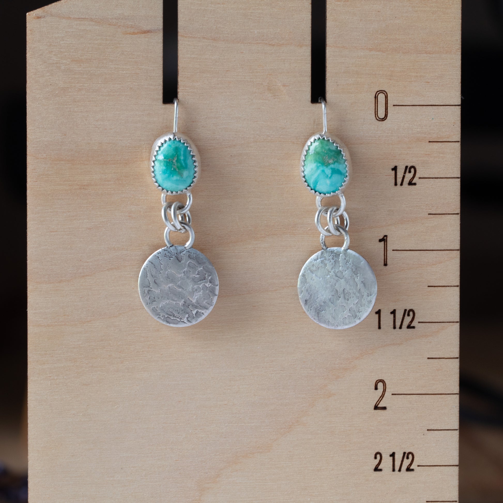 Pair of earrings with turquoise stones and silver discs on a wooden block with measurements.