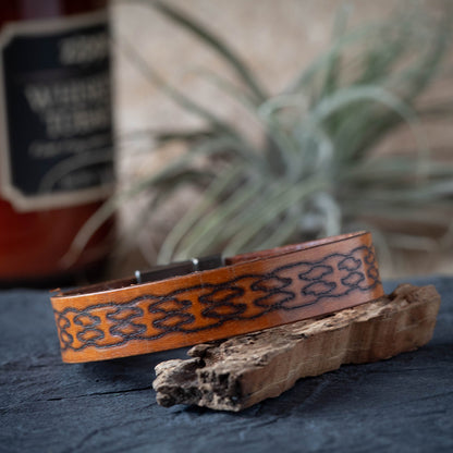 Brown leather bracelet with intricate patterns on a dark surface with a plant in the background