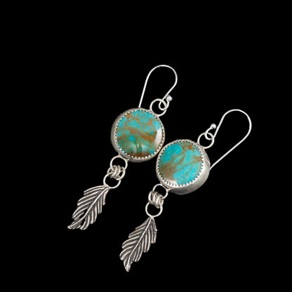 Sterling silver earrings with round bezel-set turquoise stones and dangling feather charms, handcrafted Southwestern-inspired design by Divella Designs – shown on black background.