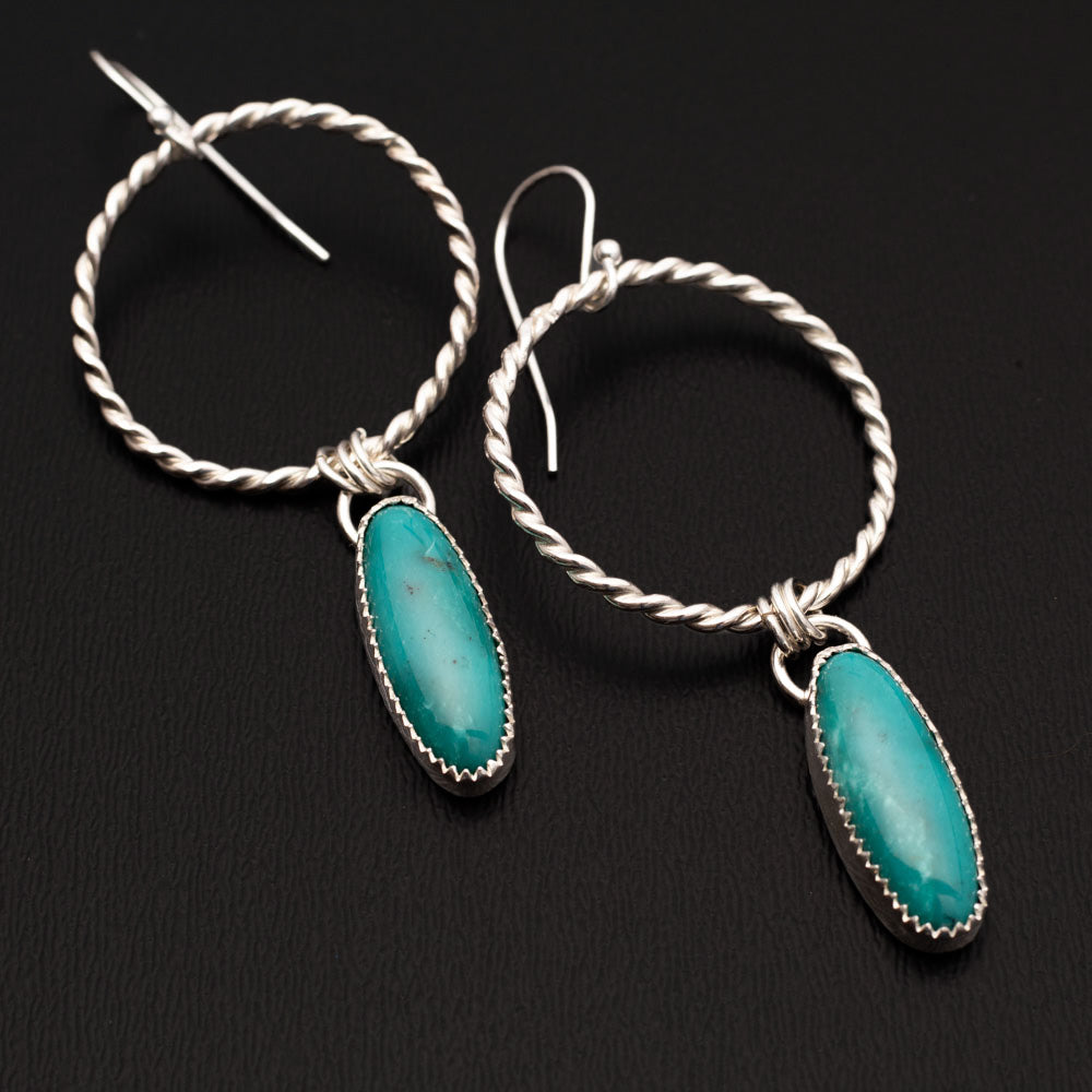 Sterling silver hoop earrings featuring oval turquoise stones on one side and hand-textured silver backs, with front and back of earrings shown on a black background.