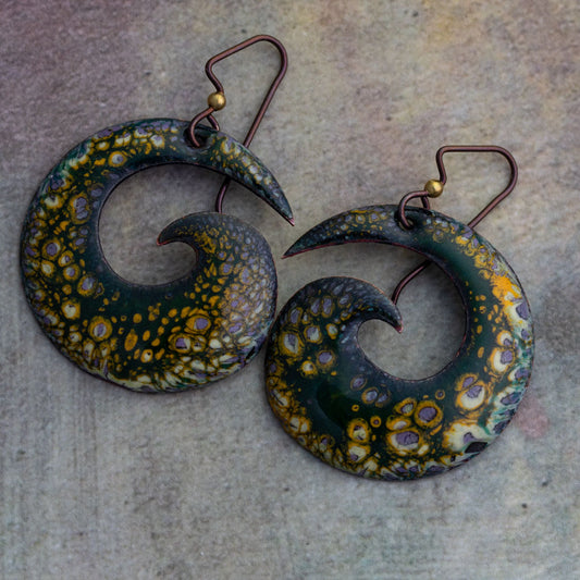 Artisan enamel spiral earrings in rich forest green with golden and violet organic patterning, crafted with niobium ear wires, photographed on a natural stone background.