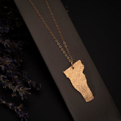 Brass Vermont-shaped pendant with embossed leaves on a gold-filled chain, displayed on a jewelry stond with sprigs of lavender.