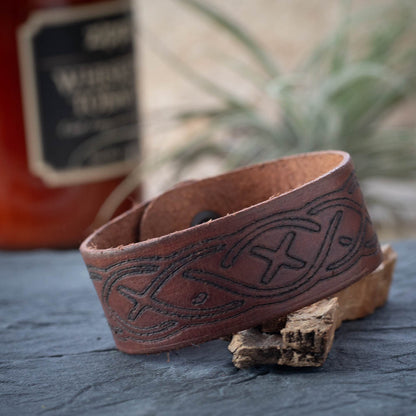 Brown leather bracelet with embossed design on a wooden surface with a blurred bottle in the background