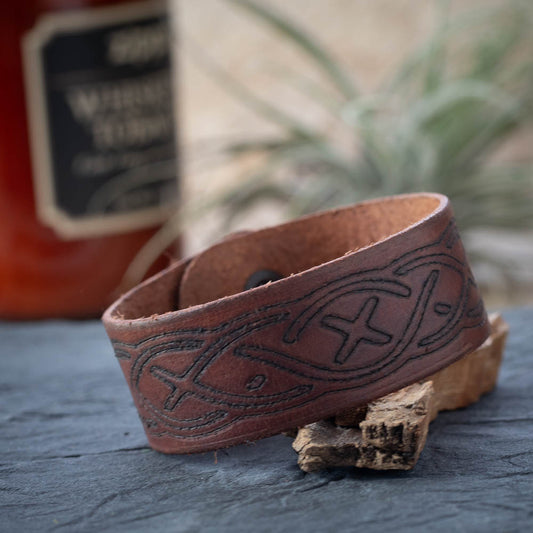 Brown leather bracelet with embossed design on a wooden surface with a blurred bottle in the background