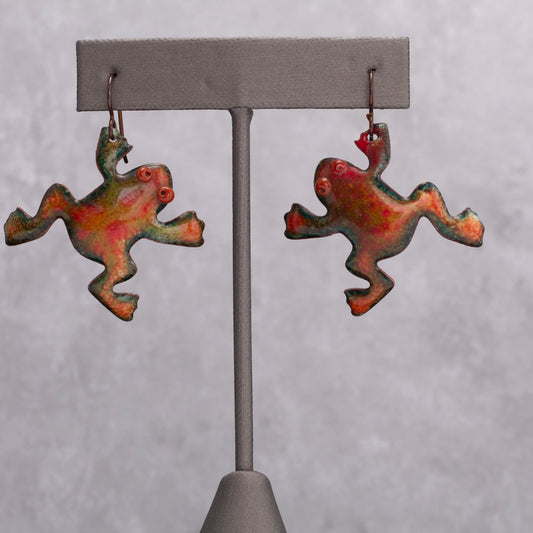 Colorful enamel frog-shaped earrings with playful red spiral eyes, displayed on a gray stand against a soft gray background.