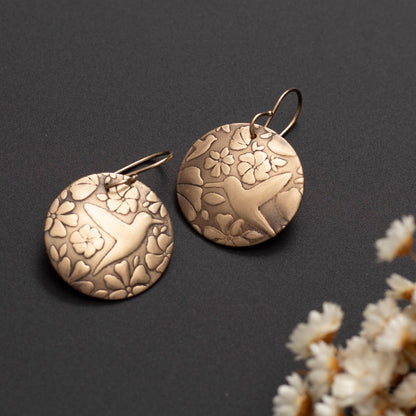 Etched brass hummingbird and floral motif earrings with gold-filled ear wires, displayed on dark background with white dried flowers.