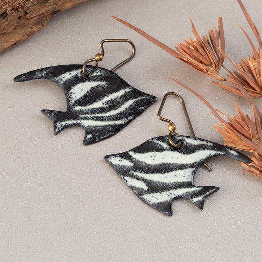 Handmade enamel fish-shaped earrings in bold white and black stripes, styled with driftwood and natural textures – artisan jewelry by Divella Designs