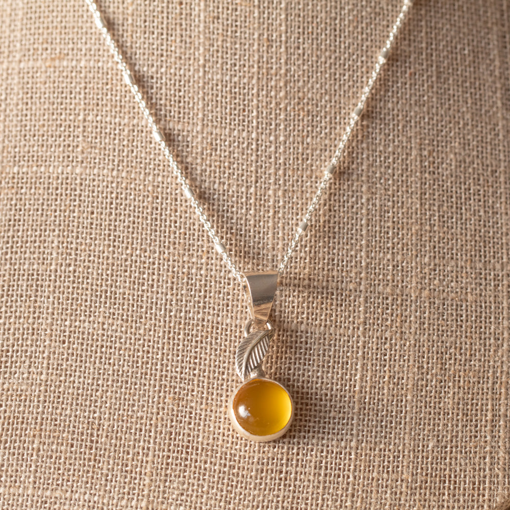 A silver pendant necklace with a leaf-shaped charm and a round yellow chalcedony cabochon stone, displayed on a piece of linen