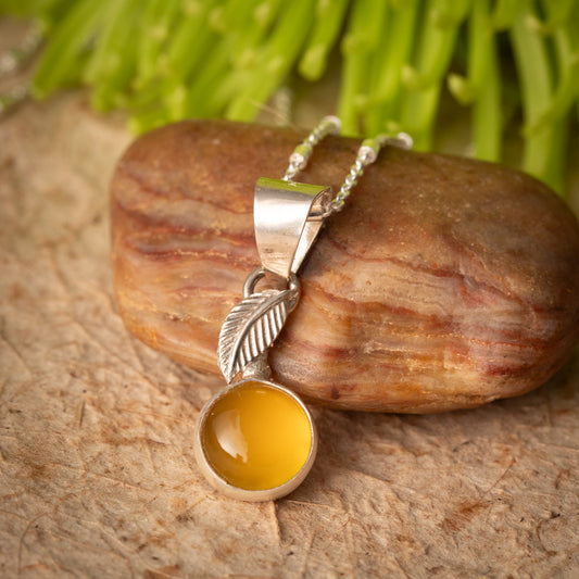 A silver pendant necklace with a leaf-shaped charm and a round yellow chalcedony cabochon stone, displayed on a brown rock with green leaves in the background.