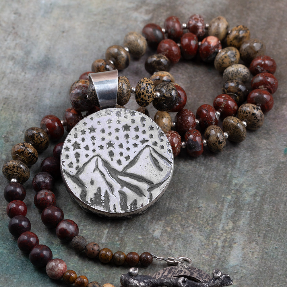 Back of a handmade sterling silver pendant featuring an engraved mountain landscape under a star-filled sky, paired with a jasper beaded necklace.
