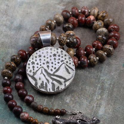 Back of a handmade sterling silver pendant featuring an engraved mountain landscape under a star-filled sky, paired with a jasper beaded necklace.