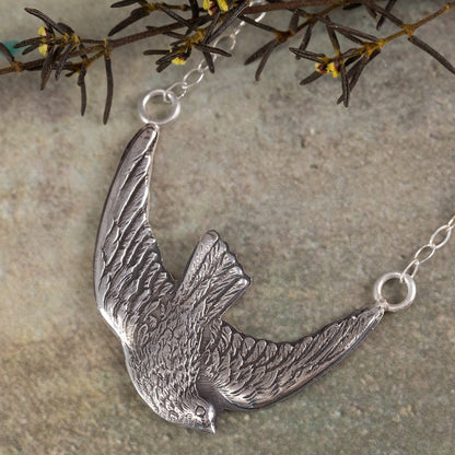 Sterling silver bird in flight pendant necklace with detailed feather texture, displayed on slate background with sprigs of greenery.