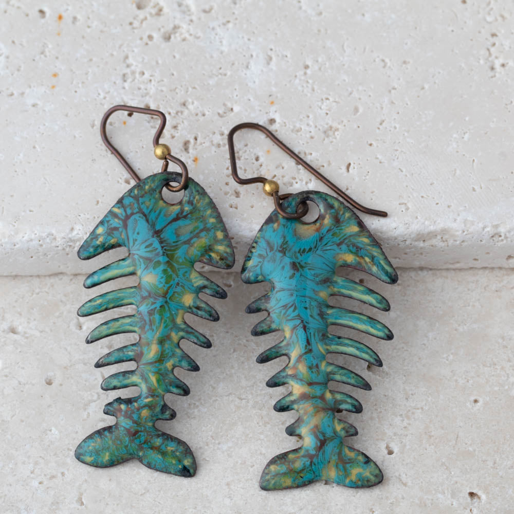 Pair of fish-shaped earrings with a textured blue and green finish on a light stone background.