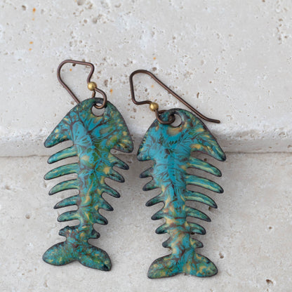 Pair of fish-shaped earrings with a textured blue and green finish on a light stone background.