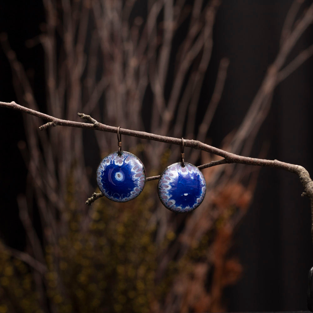 Handmade blue and purple enamel earrings photographed on a branch with  woodsy bouquet in the background.