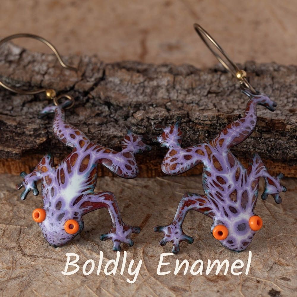 Frog-shaped earrings with purple and brown pattern on a wooden surface, featuring the brand 'Boldly Enamel'.