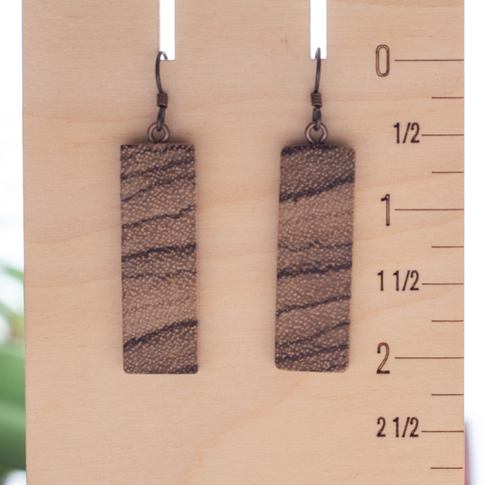 A pair of rectangular drop earrings with a textured brown and beige wood-grain pattern, hanging from dark metal hooks on a light wood display block with a size ruler printed on the side.