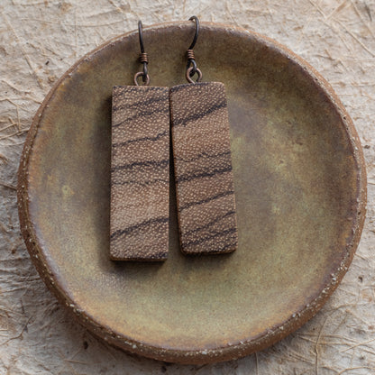 A pair of rectangular drop earrings with a textured brown and beige wood-grain pattern, hanging from niobium fishhook wires on a rustic ceramic dish.