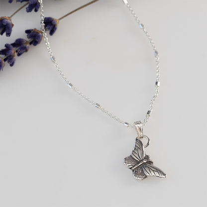 Delicate sterling silver butterfly pendant necklace with fine wing detailing, displayed on a white background with lilac sprigs.