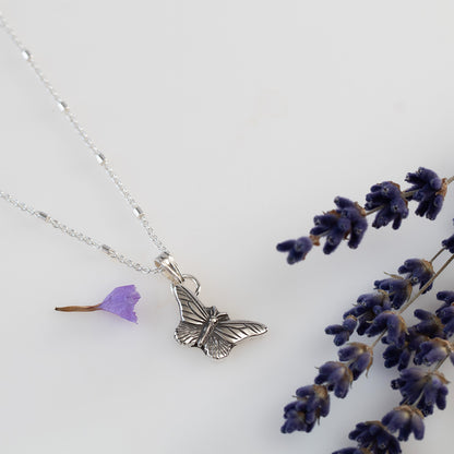 Delicate sterling silver butterfly pendant necklace with fine wing detailing, displayed on a white background with scattered purple flower petals and lilac.