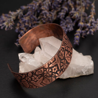 Etched copper cuff bracelet featuring a hummingbird pattern, displayed on a crystal with dried lavender on a dark background.