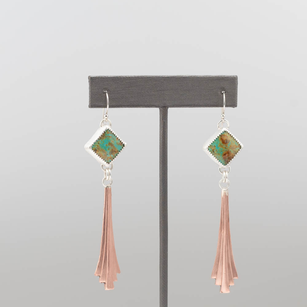 Pair of earrings with green gemstones and copper tassels on a gray stand.
