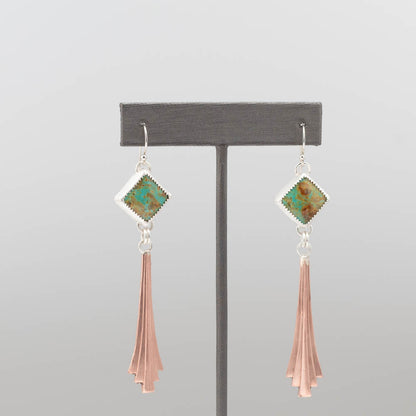 Pair of earrings with green gemstones and copper tassels on a gray stand.