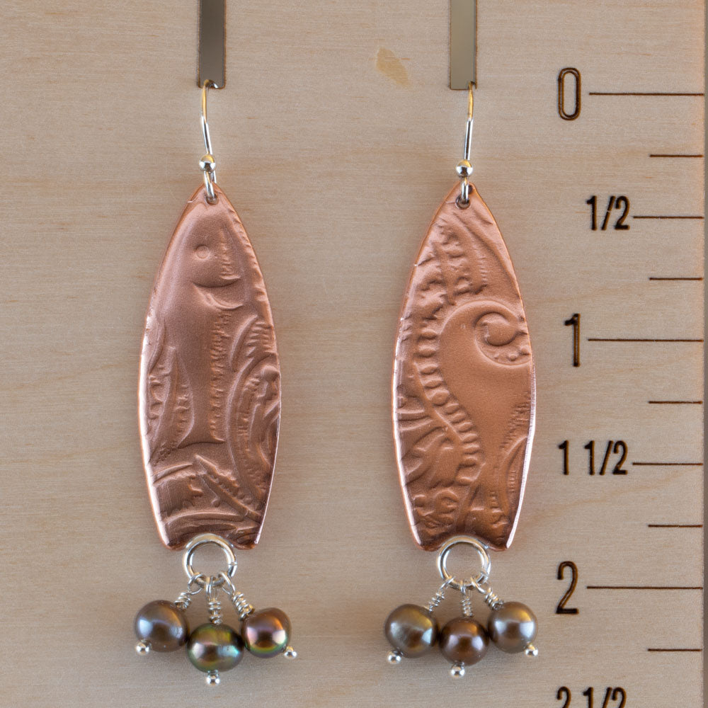 "Handcrafted copper earrings with surfboard shape and embossed pattern, accented with dangling dark freshwater pearls – Surf’s Up artisan design by Divella Designs, shown on a wooden ruler for scale.