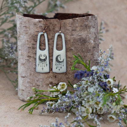 Boho-style enamel earrings in creamy white with black celestial crescent moon and star designs, handcrafted rectangular dangles with cutout tops. Displayed on birch bark with wildflowers – artisan jewelry by Divella Designs.