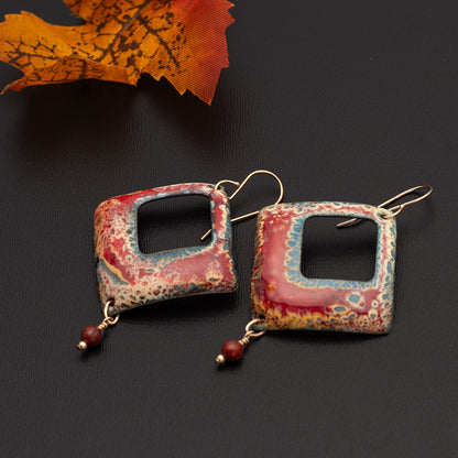 Square-shaped enamel earrings in fiery red, gold, and blue tones with center cut-outs and small red bead dangles, displayed on a dark surface with a fall leaf accents.