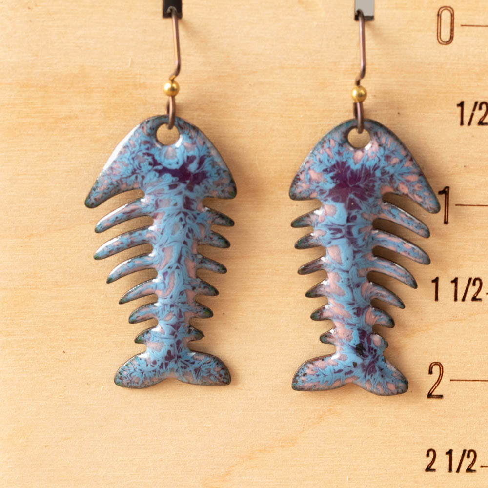 Blue and purple fish bone-shaped earrings on a wooden measuring scale.