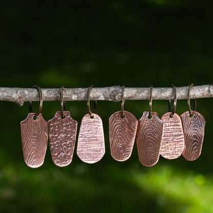 Handcrafted copper keychains with assorted embossed nature patterns hanging on a branch, artisan-made with rustic textures.