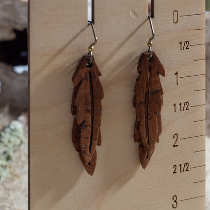 Two brown feather-shaped earrings hanging on a wooden display block with measurement markings.