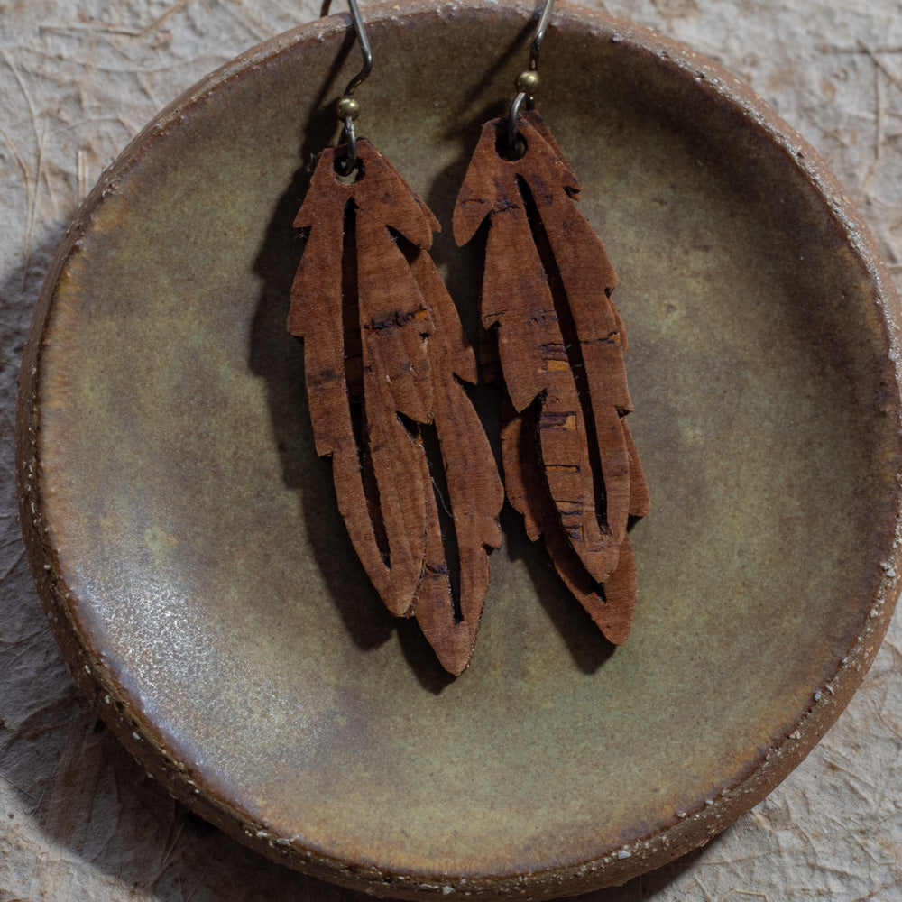 A pair of brown faux cork feather drop earrings with niobium fishhook wires, displayed on a rustic, round, textured brown ceramic dish.
