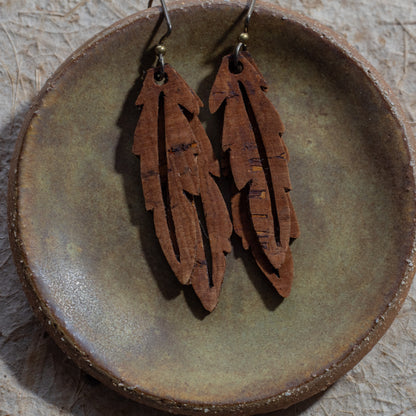 A pair of brown faux cork feather drop earrings with niobium fishhook wires, displayed on a rustic, round, textured brown ceramic dish.