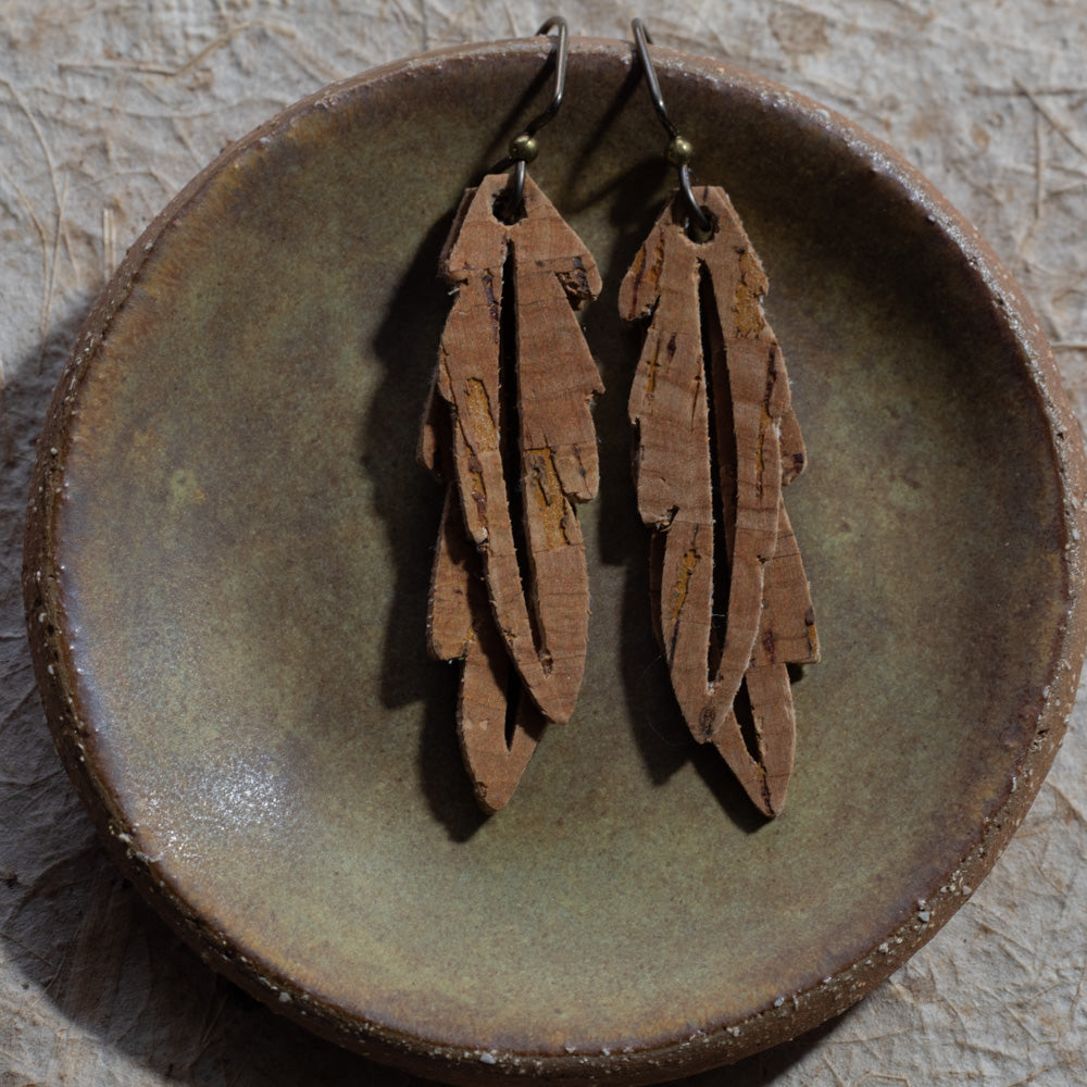 A pair of brown faux cork feather drop earrings with niobium fishhook wires, displayed on a rustic, round, textured brown ceramic dish.