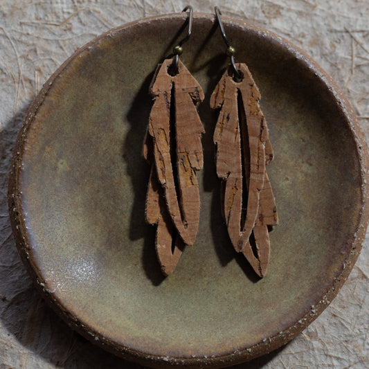 A pair of brown faux cork feather drop earrings with niobium fishhook wires, displayed on a rustic, round, textured brown ceramic dish.