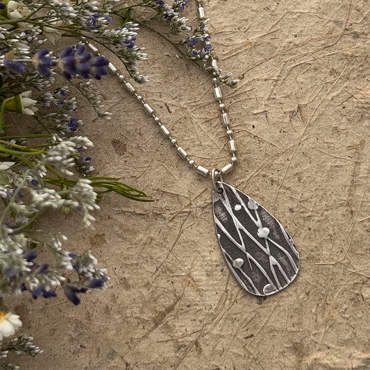 Handcrafted fine silver teardrop pendant with a raindrop and branch texture, hanging from a beaded silver chain, displayed on a natural handmade textured surface with dried lavender and white wildflowers.