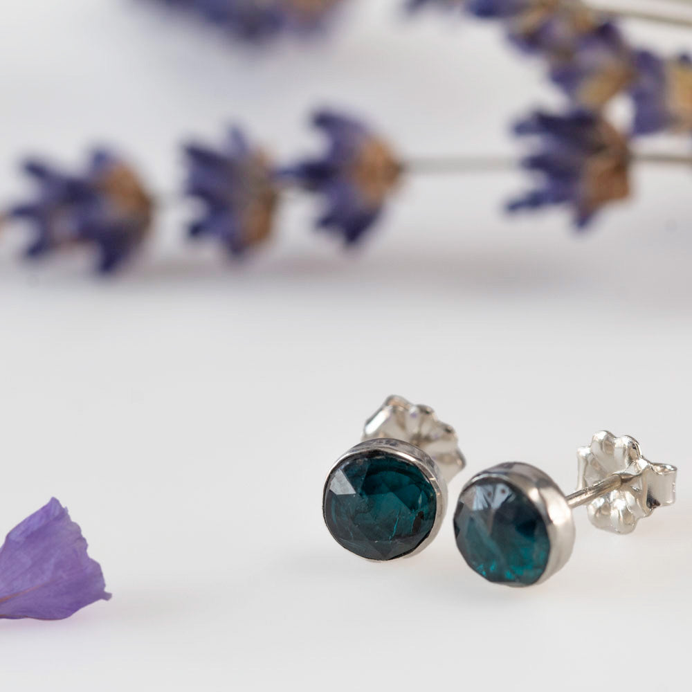 Sterling silver stud earrings featuring faceted teal kyanite gemstones, photographed on a white background with soft lilac accents.