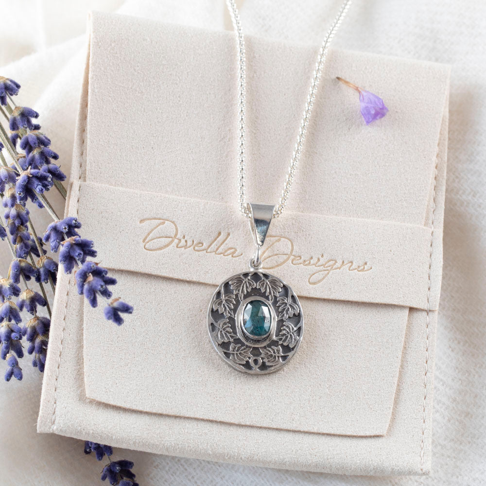 Sterling silver pendant necklace with an ornate leaf design surrounding an oval blue-green kyanite stone, shown on a branded pouch on a  white background with scattered purple flower petals