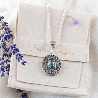 Sterling silver pendant necklace with an ornate leaf design surrounding an oval blue-green kyanite stone, shown on a branded pouch on a  white background with scattered purple flower petals