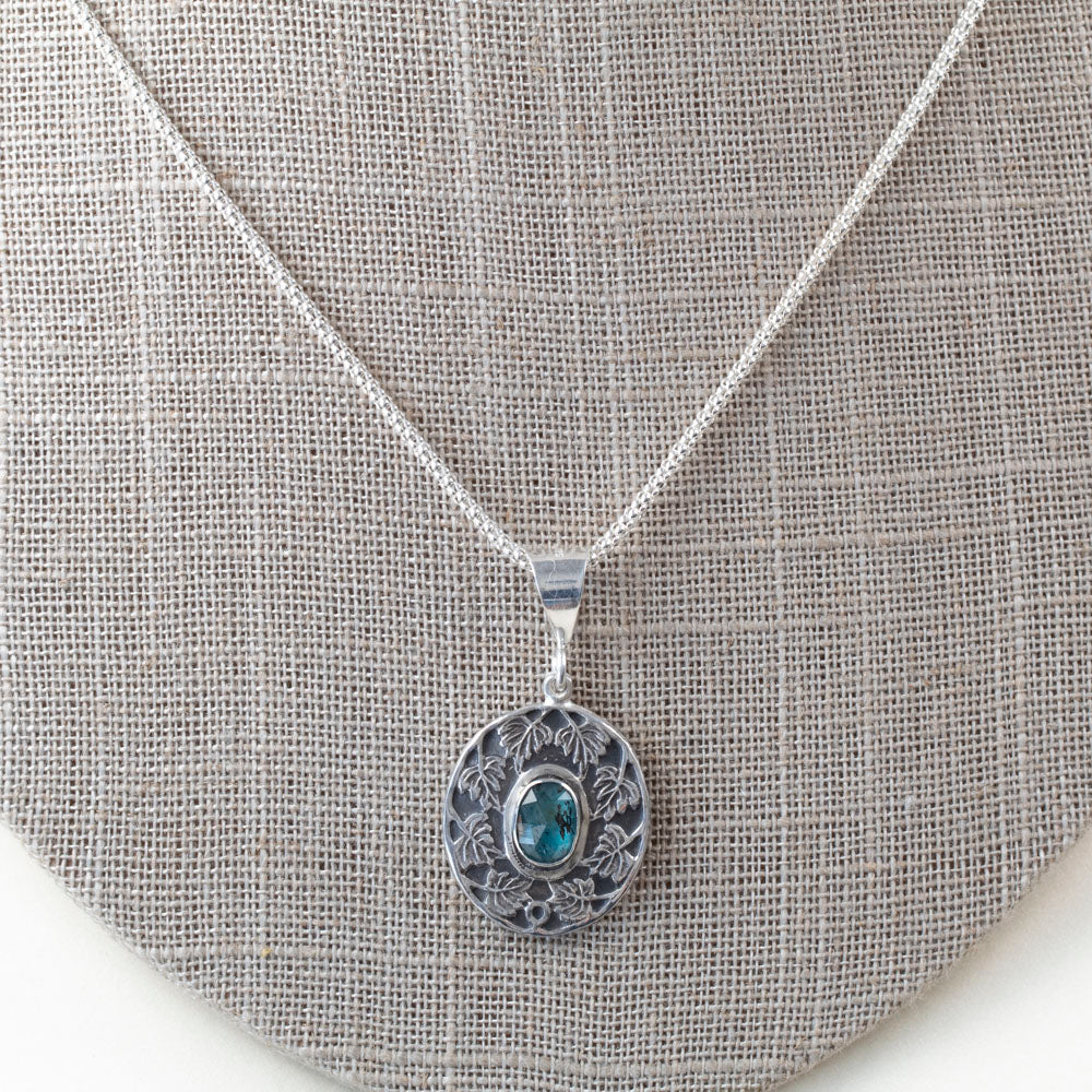 Sterling silver pendant necklace with an ornate leaf design surrounding an oval blue-green kyanite stone, shown on a gray but on a white background.