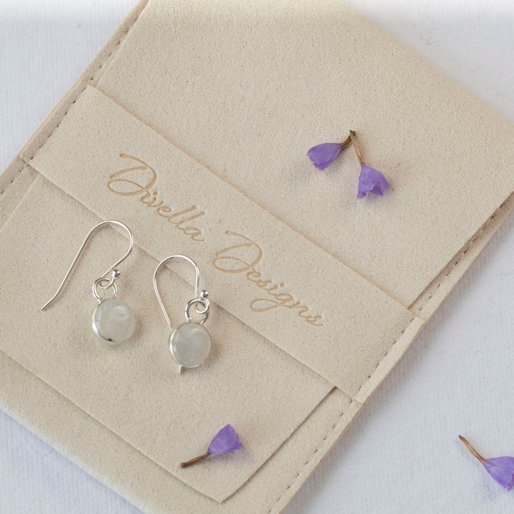 Delicate moonstone drop earrings in sterling silver with round cabochon stones, simple handmade dangles displayed with scattered purple flower petals on a branded pouch.