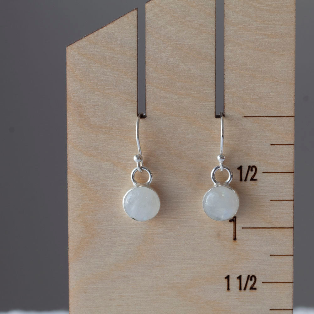 Delicate moonstone drop earrings in sterling silver with round cabochon stones, simple handmade dangles displayed with scattered purple flower petals on measurement card.