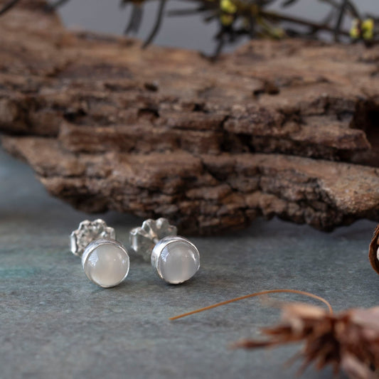 Sterling silver moonstone stud earrings with smooth round cabochons, placed on a slate-colored surface with natural bark and dried botanical elements in the background.