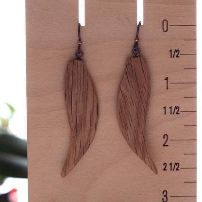A pair of wooden drop earrings with a natural wood grain finish and dark metal hooks, displayed on a wooden block with a ruler scale on the right side.
