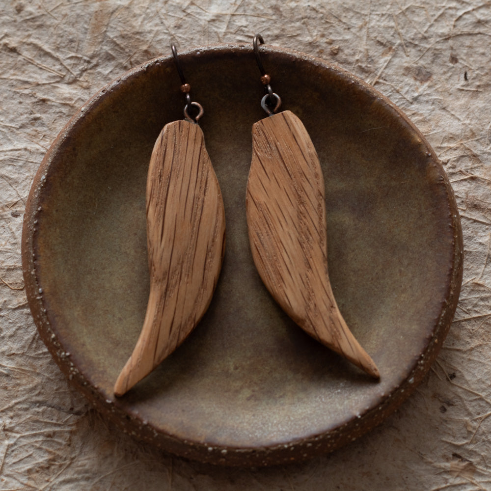 A pair of wooden dangle earrings with a natural wood grain finish, displayed on a rustic ceramic dish over a textured beige surface.