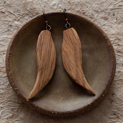 A pair of wooden dangle earrings with a natural wood grain finish, displayed on a rustic ceramic dish over a textured beige surface.