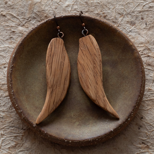 A pair of wooden dangle earrings with a natural wood grain finish, displayed on a rustic ceramic dish over a textured beige surface.